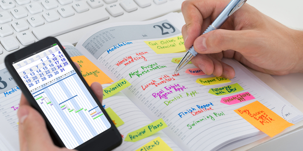 Una mano de persona escribiendo tareas en una agenda de papel mientras sujeta un móvil (abierto y mostrando un calendario digital).
